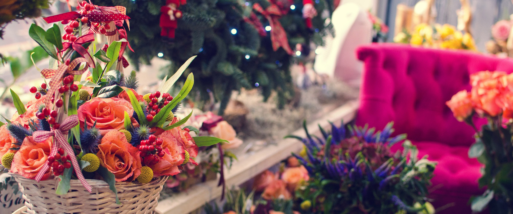 A wicker basket filled with orange roses, green leaves, and red berries sits on a table in a festive, decorated room with a pink tufted sofa and a Christmas tree.