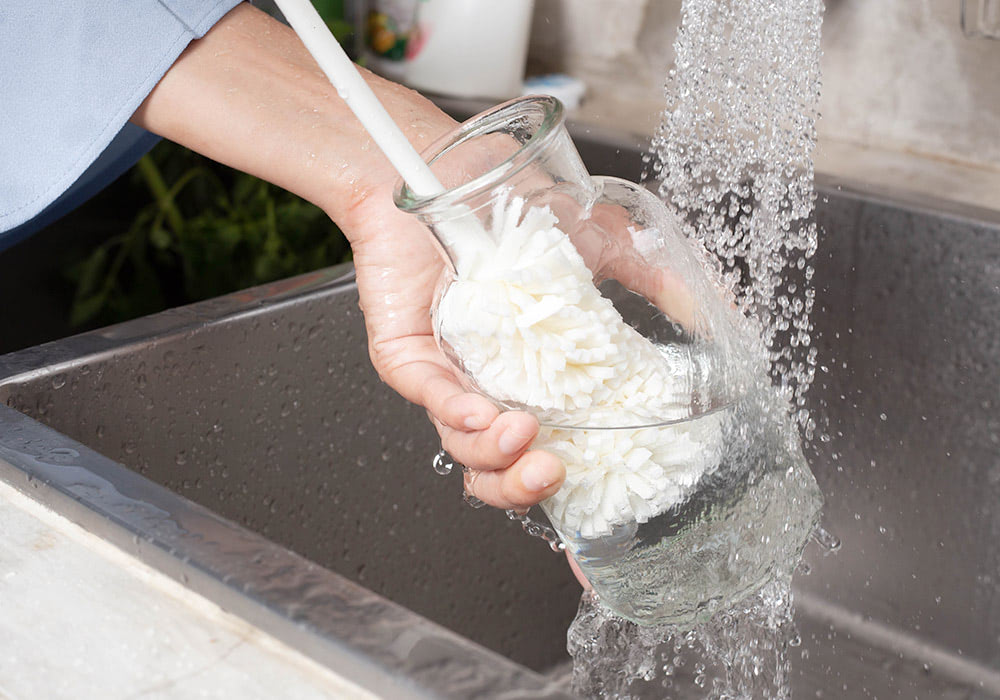 A hand holding a clear pitcher scrubs its interior with a white brush while being washed under running water in a stainless steel sink.