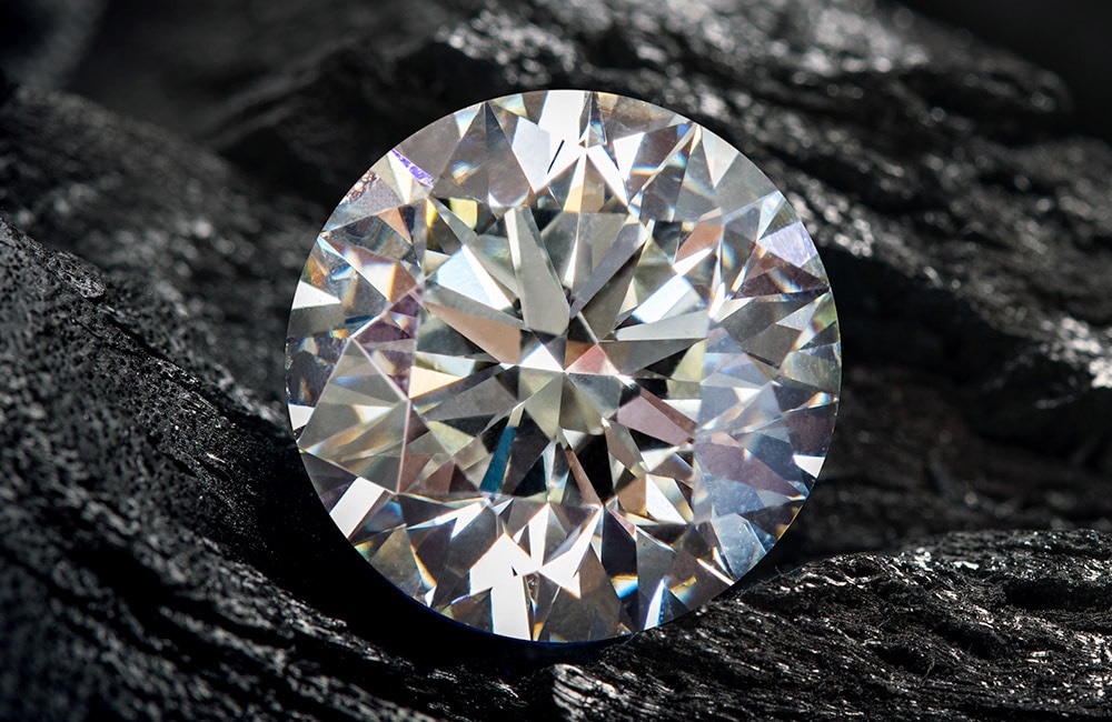 A brilliant-cut diamond resting on a rough, dark-textured surface, reflecting a spectrum of light, highlighting its facets and clarity amidst the contrast of its rugged backdrop.