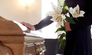 A person in black holds white lilies while placing a hand on a wooden coffin in a softly lit, indoor setting.